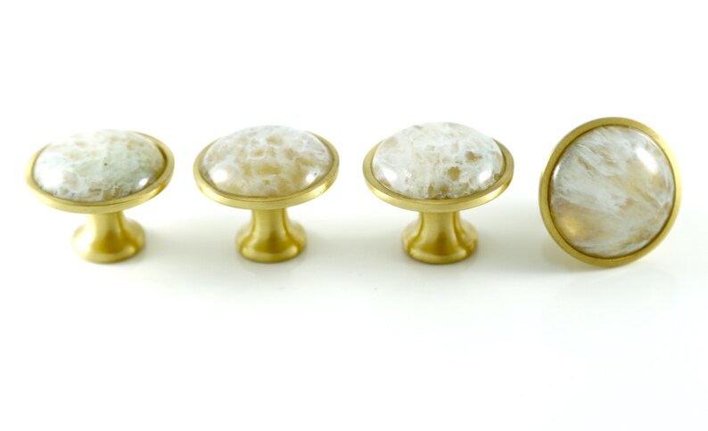 K&ouml;nnte beinhalten: Vier goldene Schrankkn&ouml;pfe mit wei&szlig;en Achat-Steinoberfl&auml;chen. Die Kn&ouml;pfe sind rund und haben eine glatte, polierte Oberfl&auml;che.