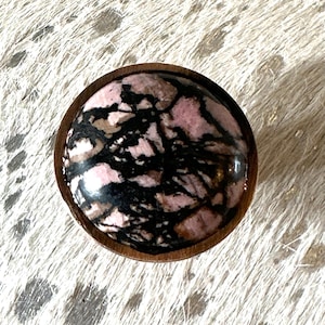 Op de afbeelding: Een ronde, houten knop met een roze en zwarte marmeren steeninleg. De steen heeft een draaiende patroon van roze en zwart.