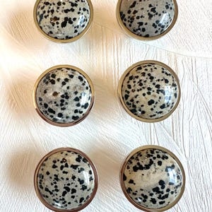 Könnte beinhalten: Sechs runde Schrankknöpfe mit einem gesprenkelten, hellgrauen und schwarzen Stein in einem goldfarbenen Metallrahmen. Die Knöpfe sind in zwei Dreierreihen auf einem weißen, strukturierten Hintergrund angeordnet.