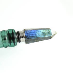 Puede incluir: Una botella de vidrio verde con un tap&oacute;n de cristal azul iridiscente y plateado.