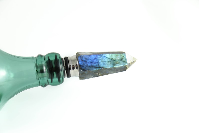 Puede incluir: Una botella de vidrio verde con un tap&oacute;n de cristal azul iridiscente y plateado.