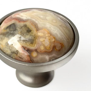 May include: A round, silver cabinet knob with a natural agate stone inlay. The stone has a swirling pattern of white, brown, and pink.