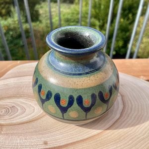 Könnte beinhalten: Eine runde Keramikvase mit gesprenkelter grüner und beiger Glasur. Der Körper ist mit einem sich wiederholenden Muster aus dunkelblauen, Y-förmigen Motiven und kleinen orangefarbenen Punkten verziert. Der Rand hat ein dunkelblaues Band.