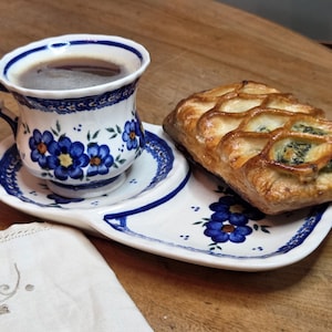 Puede incluir: Un plato de cerámica blanca para aperitivos con portavasos integrado presenta motivos florales azules. Una taza a juego llena de café oscuro se asienta en el soporte, y un hojaldre dorado con rejilla relleno de espinacas descansa sobre el plato.