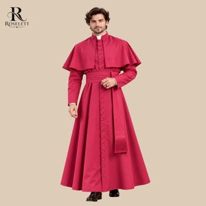 May include: A vibrant magenta cassock featuring a shoulder cape, a high collar with a white tab insert, and a long row of matching fabric buttons down the front. A wide sash with a fringed end is tied at the waist. The Roselett logo is visible.