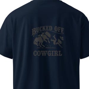 May include: A dark navy blue short-sleeved t-shirt featuring a dark grey graphic on the back. The design depicts a bucking horse and a falling figure with a hat, accompanied by the text BUCKED OFF COWGIRL in a bold, western-style font.