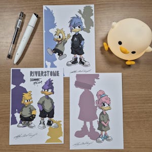 Pode incluir: Três impressões de arte retangulares com personagens de patos de desenhos animados, uma caneta de tinta preta, um marcador branco e uma lâmpada amarela em forma de pintinho. Uma impressão exibe o texto RIVERSTONE SCHOOL PRIDE.