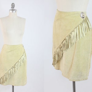 May include: A light beige suede skirt with a diagonal fringe trim and a silver button detail. The skirt is a wrap style and has a mini length.