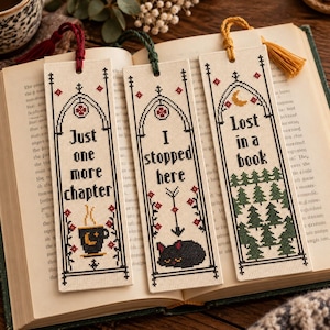 Pode incluir: Três marcadores de página retangulares em ponto cruz sobre um livro. O primeiro tem uma borla vermelha e diz Just one more chapter com uma chávena. O segundo tem uma borla verde e diz I stopped here com um gato. O terceiro tem uma borla amarela e diz Lost in a book com árvores.
