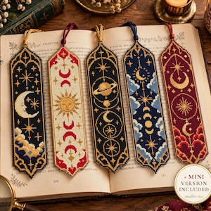 May include: Five rectangular cross-stitched bookmarks with pointed ends and tassels, featuring celestial designs including moons, suns, stars, planets, and clouds in black, cream, blue, and red thread. A circular label reads + MINI VERSION INCLUDED.
