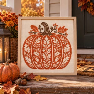 May include: A square framed cross-stitch embroidery featuring a large orange pumpkin design. The pumpkin is decorated with white oak leaf and acorn patterns, topped with a brown stem and small sprigs of berries, all set against a cream fabric.