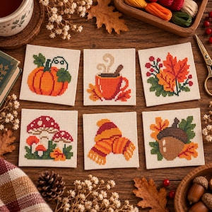 May include: Six square cross-stitch fabric patches featuring autumn designs: an orange pumpkin, a steaming mug, colourful fall leaves, red and white mushrooms, a striped knit hat and scarf, and a brown acorn with leaves on a cream background.