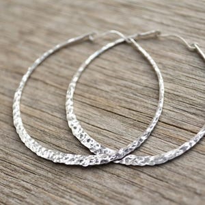 May include: A pair of large, circular hoop earrings crafted from silver-toned metal. The surface of each hoop features a distinct, irregular hammered texture that catches the light, creating a reflective and uneven finish along the entire circumference.