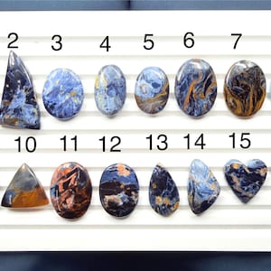May include: Sixteen polished blue and brown gemstones arranged in two rows on a white display board. Each stone is numbered from 1 to 16 and features unique swirling patterns of deep blue, black, and golden-brown colors in various shapes.