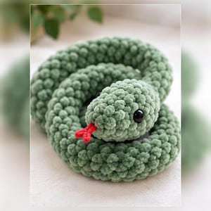 Può includere: Un serpente di peluche arrotolato realizzato in filato testurizzato verde salvia. Il peluche presenta una testa arrotondata con occhi in plastica nera liscia e una piccola lingua biforcuta in filato rosso vivo che sporge.