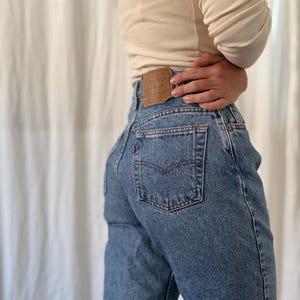 Puede incluir: Vaqueros de mezclilla azul de talle alto con una etiqueta de cuero color canela en la cintura trasera, una pequeña etiqueta roja en el bolsillo trasero y remaches clásicos de color cobre. Los vaqueros se combinan con una blusa de punto acanalado color crema.