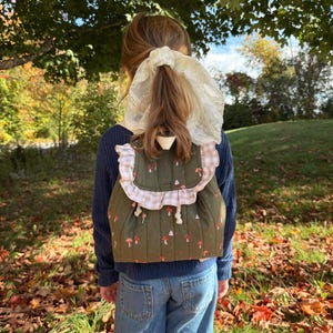 May include: A quilted olive green backpack featuring a repeating pattern of small red and white mushrooms. The bag has a light beige and white gingham ruffled trim along the top flap and white drawstring cords for closure, worn over a navy sweater.