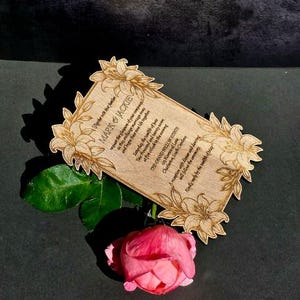 May include: A rectangular wooden wedding invitation card with laser-engraved lily floral corners and text reading Mark & Jackie, The Grand Willow Estate, Charleston, South Carolina. A single pink rose with green leaves rests beneath the card.
