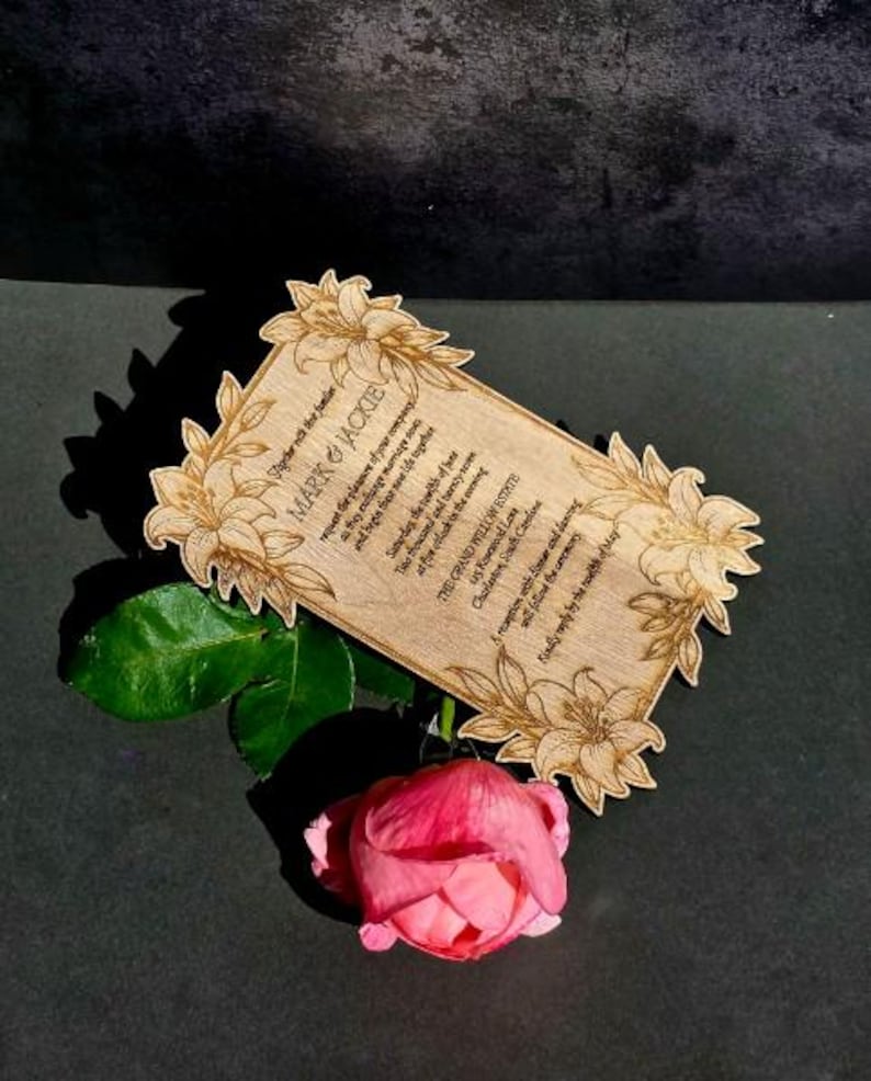 May include: A rectangular wooden wedding invitation card with laser-engraved lily floral corners and text reading Mark & Jackie, The Grand Willow Estate, Charleston, South Carolina. A single pink rose with green leaves rests beneath the card.