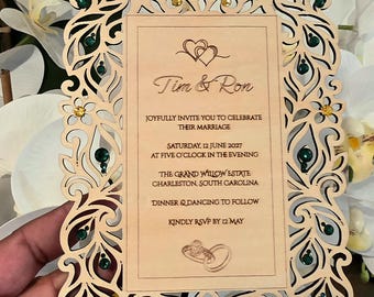 May include: A rectangular wooden wedding invitation featuring intricate laser-cut peacock motifs along the border, accented with small green and yellow rhinestones. The text reads: Tim & Ron, joyfully invite you to celebrate their marriage, Saturday, 12 June 2027, at five o'clock in the evening, The Grand Willow Estate, Charleston, South Carolina, dinner & dancing to follow, kindly RSVP by 12 May.
