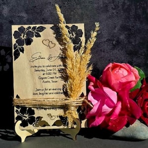 Peut inclure: Une carte d'invitation de mariage rectangulaire en bois avec des illustrations d'hibiscus noirs, deux petits c&oelig;urs et du texte imprim&eacute;. La carte est attach&eacute;e avec de la ficelle, d&eacute;cor&eacute;e d'&eacute;pis de bl&eacute; s&eacute;ch&eacute;s et pos&eacute;e sur un petit support en bois.