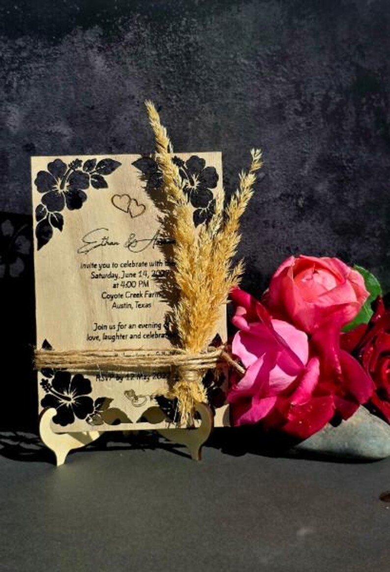 Peut inclure: Une carte d'invitation de mariage rectangulaire en bois avec des illustrations d'hibiscus noirs, deux petits c&oelig;urs et du texte imprim&eacute;. La carte est attach&eacute;e avec de la ficelle, d&eacute;cor&eacute;e d'&eacute;pis de bl&eacute; s&eacute;ch&eacute;s et pos&eacute;e sur un petit support en bois.