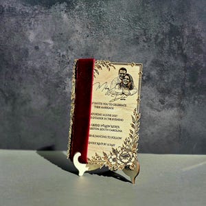 May include: A rectangular wooden wedding invitation featuring a laser-engraved portrait of a couple, floral borders, and text reading Mike & Sadie, invite you to celebrate their marriage, Saturday, 12 June 2027, at 5 o'clock in the evening, Grand Willow Estate, Charleston, South Carolina, dinner & dancing to follow, kindly RSVP by 12 May, accented with a deep red velvet ribbon.