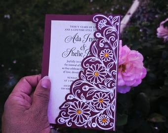 May include: A maroon and white invitation card featuring an intricate laser-cut floral overlay on the right side. The design includes white daisy-like flowers with small amber-colored rhinestones at their centers. The text reads Thirty Years of Love and a Lifetime Still to Go, Ada Ing & Shelie J, Joyfully invite you to the renewal of their vows as they celebrate thirty years of love, devotion, and friendship.