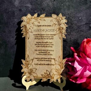May include: A rectangular wooden wedding invitation plaque with laser-engraved lily floral borders. The text reads: Together with their families MARK & JACKIE request the pleasure of your company as they exchange marriage vows and begin their new life together. Saturday the twelfth of June Two thousand and twenty-seven at five o'clock in the evening THE GRAND WILLOW ESTATE 125 Rosewood Lane Charleston, South Carolina. A reception with dinner and dancing will follow the ceremony. Kindly reply by the twelfth of May.