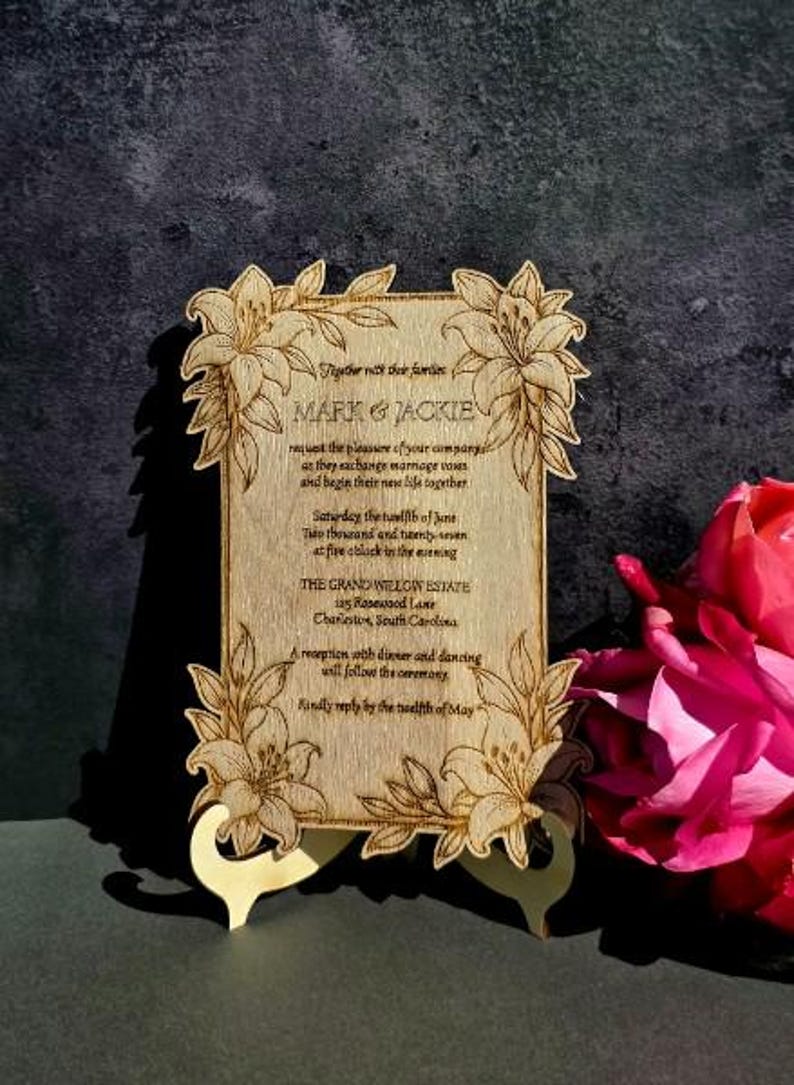 May include: A rectangular wooden wedding invitation plaque with laser-engraved lily floral borders. The text reads: Together with their families MARK & JACKIE request the pleasure of your company as they exchange marriage vows and begin their new life together. Saturday the twelfth of June Two thousand and twenty-seven at five o'clock in the evening THE GRAND WILLOW ESTATE 125 Rosewood Lane Charleston, South Carolina. A reception with dinner and dancing will follow the ceremony. Kindly reply by the twelfth of May.