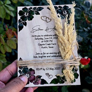 Peut inclure: Une carte d'invitation de mariage rectangulaire en bois avec des illustrations d'hibiscus noirs, deux c&oelig;urs et deux anneaux. Le texte indique Ethan & A, samedi 14 juin 2027, 16h00, Coyote Creek Farm, Austin, Texas, et RSVP avant le 12 mai 2027.