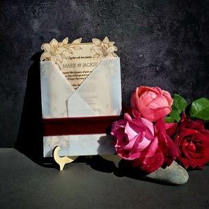 May include: A rectangular wooden wedding invitation with laser-cut lily floral details at the top, enclosed in a translucent vellum envelope wrap secured by a dark red velvet ribbon, featuring the text Mark & Jackie.
