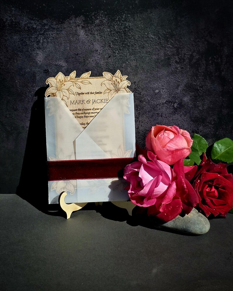 May include: A rectangular wooden wedding invitation with laser-cut lily floral details at the top, enclosed in a translucent vellum envelope wrap secured by a dark red velvet ribbon, featuring the text Mark & Jackie.