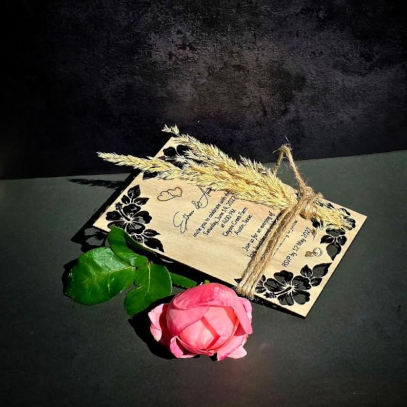 Peut inclure: Une carte d'invitation de mariage rectangulaire en bois avec des motifs d'hibiscus noirs, deux petits c&oelig;urs et du texte imprim&eacute;. La carte est attach&eacute;e avec de la ficelle de jute naturelle et orn&eacute;e d'&eacute;pis de bl&eacute; s&eacute;ch&eacute;s et d'une rose rose.
