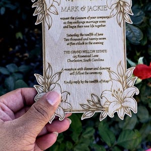 May include: A rectangular wooden wedding invitation with laser-engraved lily floral corners and text reading: Together with their families MARK & JACKIE request the pleasure of your company as they exchange marriage vows and begin their new life together.