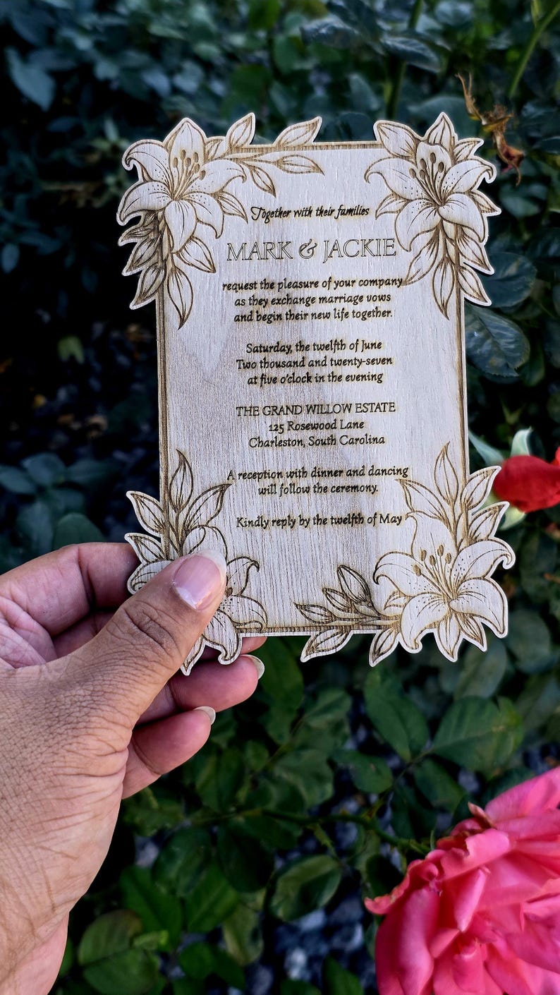 May include: A rectangular wooden wedding invitation with laser-engraved lily floral corners and text reading: Together with their families MARK & JACKIE request the pleasure of your company as they exchange marriage vows and begin their new life together.