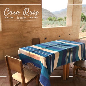 May include: A rectangular table covered with a blue, teal, and white striped textile featuring fringe edges. The table is surrounded by three wooden chairs. The text "Casa Ruiz Textiles" is displayed on the wall above the table.