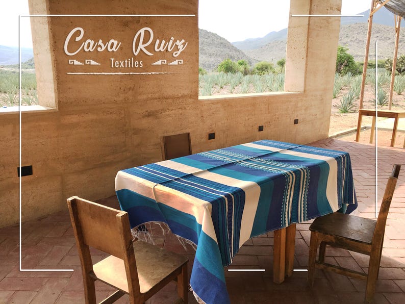 May include: A rectangular table covered with a blue, teal, and white striped textile featuring fringe edges. The table is surrounded by three wooden chairs. The text "Casa Ruiz Textiles" is displayed on the wall above the table.