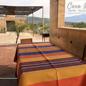 May include: A rectangular tablecloth featuring horizontal stripes in shades of orange, yellow, and blue, with a decorative woven pattern in blue and orange. The text Casa Ruiz Textiles is visible in the upper right corner of the image.