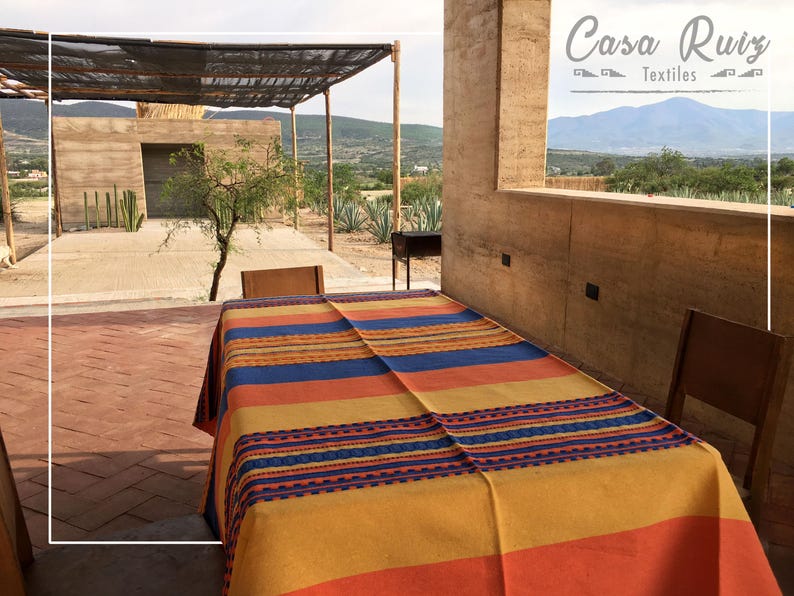 May include: A rectangular tablecloth featuring horizontal stripes in shades of orange, yellow, and blue, with a decorative woven pattern in blue and orange. The text Casa Ruiz Textiles is visible in the upper right corner of the image.
