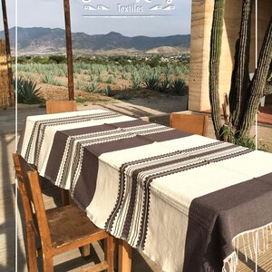 May include: A rectangular tablecloth featuring a cream-colored base with wide dark brown vertical stripes and intricate geometric border patterns. The fabric edges are finished with long, thin white fringe tassels hanging from the corners.