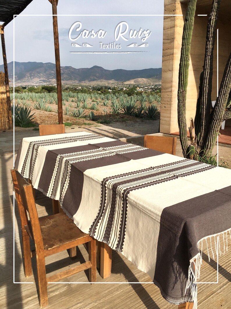 May include: A rectangular tablecloth featuring a cream-colored base with wide dark brown vertical stripes and intricate geometric border patterns. The fabric edges are finished with long, thin white fringe tassels hanging from the corners.