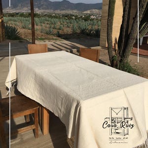 May include: A rectangular off-white tablecloth with a textured woven pattern and fringe along the edges. The fabric features a black logo printed on the corner that reads Casa Ruiz Textiles Artesanias with a graphic of a wooden loom.