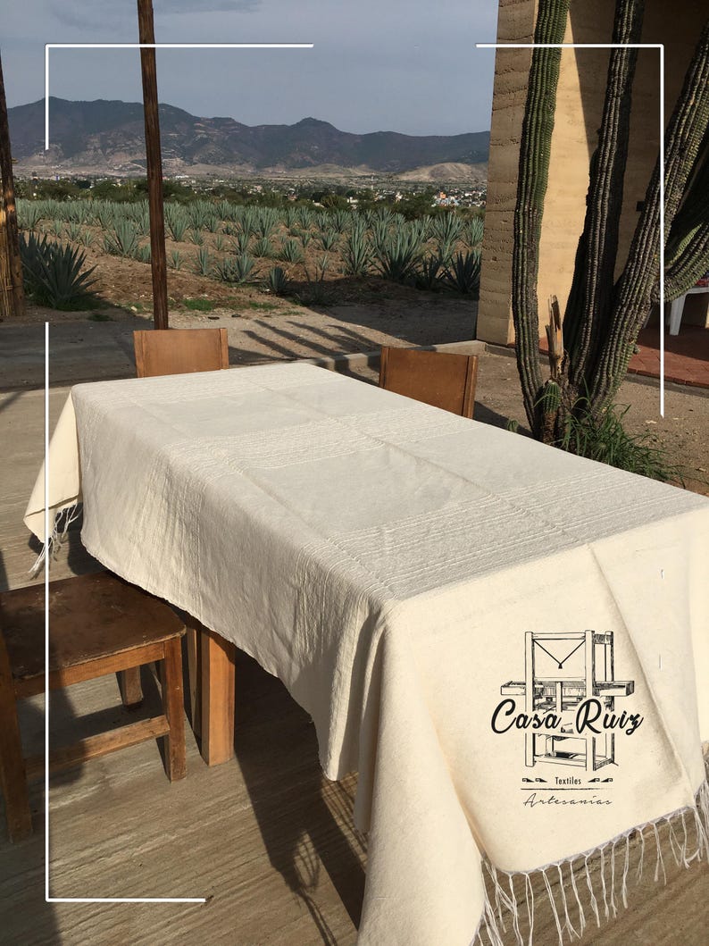 May include: A rectangular off-white tablecloth with a textured woven pattern and fringe along the edges. The fabric features a black logo printed on the corner that reads Casa Ruiz Textiles Artesanias with a graphic of a wooden loom.
