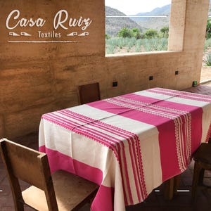 May include: A rectangular tablecloth featuring alternating wide stripes of vibrant hot pink and off-white fabric. The design includes intricate woven detailing with thin pink lines and decorative borders. The text Casa Ruiz Textiles is visible.