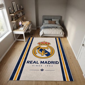 Op de afbeelding: Een rechthoekig vloerkleed met het logo van voetbalclub Real Madrid in het midden, met daaronder de tekst REAL MADRID SINCE 1902. Het kleed heeft een crèmekleurige achtergrond met verticale marineblauwe en gouden strepen aan de zijkanten.