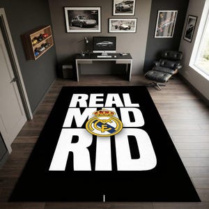 Op de afbeelding: Een rechthoekig zwart vloerkleed met de witte tekst REAL MAD RID in grote hoofdletters. Gecentreerd tussen de tekst staat het officiële embleem van voetbalclub Real Madrid, voorzien van een gouden kroon en gele en blauwe kleurdetails.