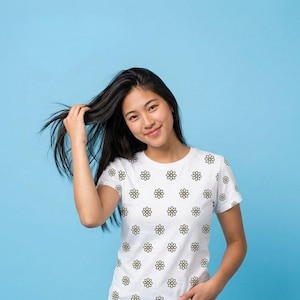 Puede incluir: Una camiseta blanca de manga corta con un patrón repetitivo de pequeñas margaritas con centros amarillos y pétalos blancos delineados en negro. La prenda cuenta con un cuello redondo y un corte estándar con un estampado floral uniforme.