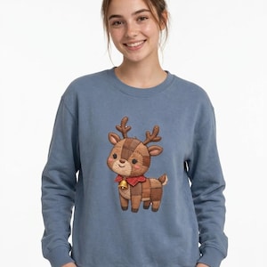 May include: A dusty blue crewneck sweatshirt featuring a central graphic of a brown patchwork-style reindeer. The reindeer has stitched detailing, a red plaid collar, and a small gold bell. The design has a soft, textured appearance.