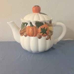 Peut inclure: Une théière en céramique blanche avec une forme côtelée rappelant une citrouille. Le corps présente un motif peint à la main de citrouilles orange, de feuilles vertes et de noix brunes. Le couvercle est surmonté d'une petite citrouille orange.